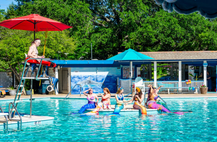 A group of people in colorful mermaid costumes sit on pool floats in a swimming pool, interacting and laughing—a playful twist on classic things to do. A lifeguard under a red umbrella watches as trees and a pool building complete the scene.