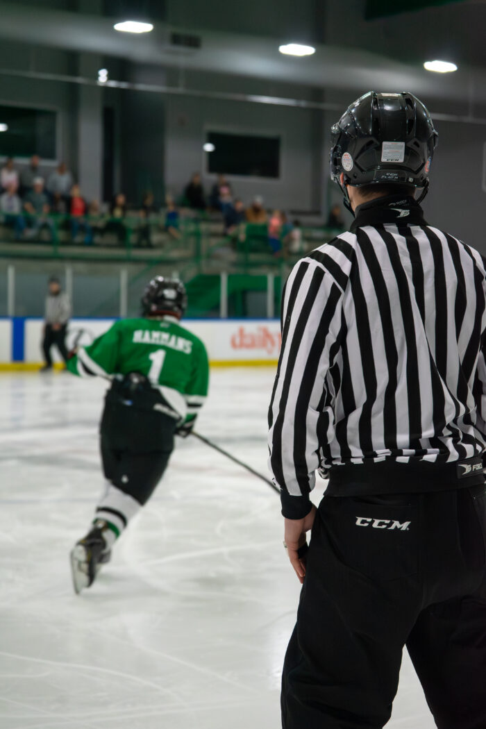 A hockey referee in black and white stripes stands on the ice, watching a player in a green jersey skate toward the rink’s far end—one of the exciting things to do for fans seated behind the glass at Children's Health StarCenter Plano