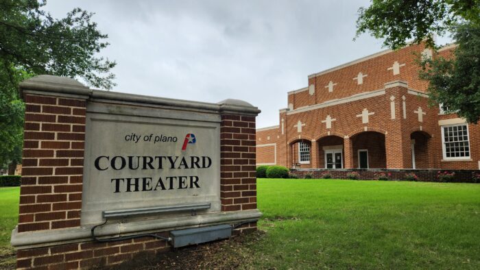 A brick sign reading City of Plano Courtyard Theater stands on a grassy lawn in front of the historic Courtyard Theater, its red brick exterior with arched doorways and white trim framed by trees beneath a cloudy sky.