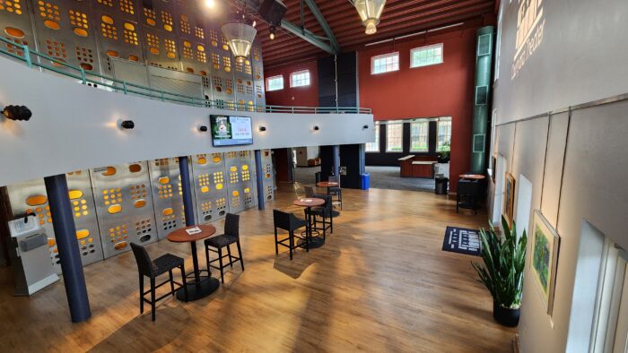 A modern, spacious Courtyard Theater lobby with wood floors, round tables and chairs, a curved balcony, decorative wall panels with light, large windows, and indoor plants. Natural light fills the room.