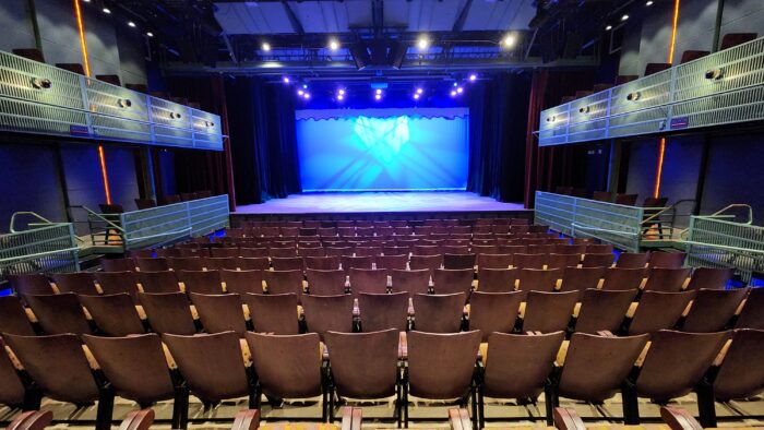 View from the back of the Courtyard Theater’s empty auditorium, with rows of brown seats, balcony seating on both sides, and a stage aglow in blue spotlights.