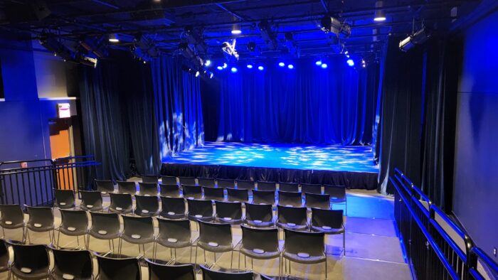 Rows of empty black chairs face a small stage, illuminated by blue spotlights, in the Cox Building Playhouse’s dark black-box theater with black curtains and ceiling-mounted stage lights.