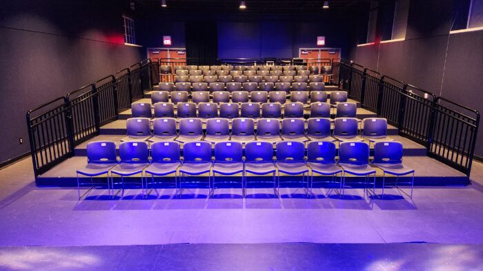 Rows of empty blue chairs are arranged in the Cox Building Playhouse auditorium, with black railings on both sides and doors at the back, all illuminated by purple and blue stage lights.