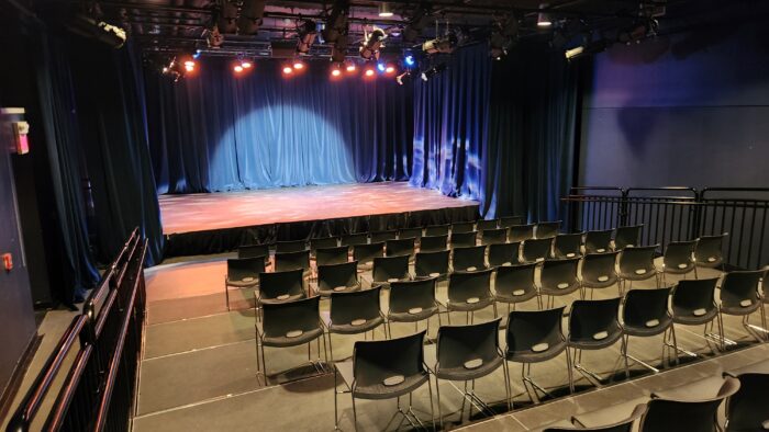 Rows of empty black chairs face a small stage with blue curtains and warm stage lighting, creating an intimate theater setting at the Cox Building Playhouse.