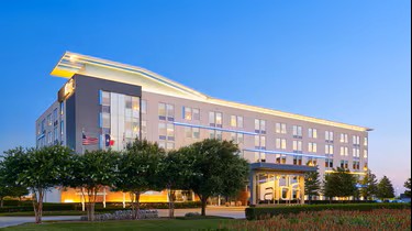 A modern four-story hotel, Aloft Plano, with large windows and a unique roof, is surrounded by trees and a well-kept lawn, shown at sunset under a clear blue sky.