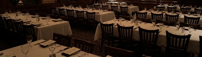 A dimly lit dining room at Bob's Steak features several tables set for an event. Each table is covered with a white tablecloth and neatly arranged with menus, glasses, and silverware. Wooden chairs surround the tables as chandeliers hang from the ceiling.
