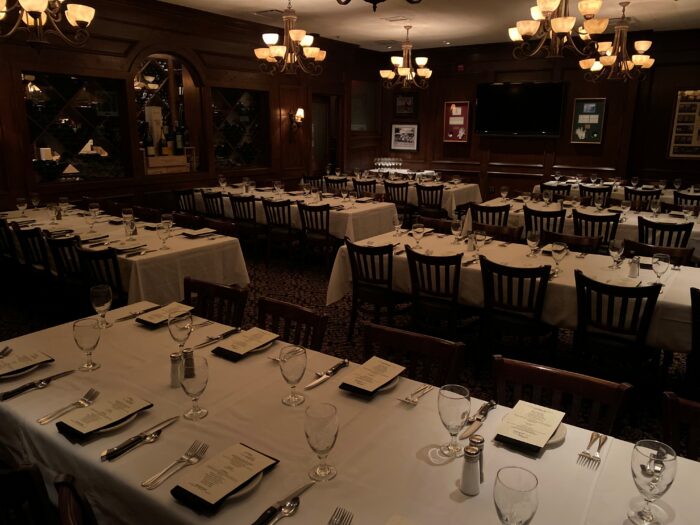 A dimly lit dining room at Bob's Steak features several tables set for an event. Each table is covered with a white tablecloth and neatly arranged with menus, glasses, and silverware. Wooden chairs surround the tables as chandeliers hang from the ceiling.