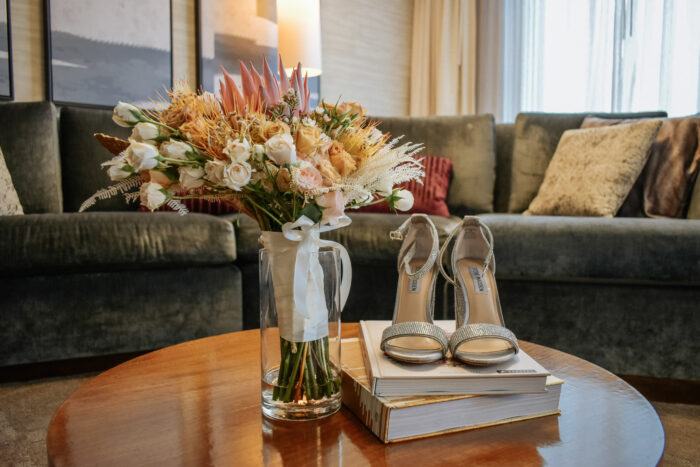 A bouquet of flowers in a glass vase and glittery high-heeled shoes sit on two books atop a round wooden table, bringing chic style to a cozy living room—just the kind of elegance you’d find at Renaissance Dallas.