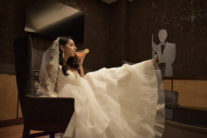 A bride in a white gown and veil relaxes in a chair with her feet up, sipping a cocktail with a lemon slice at Renaissance Dallas. The dimly lit room features a cutout figure of a man in a tuxedo on the wall.