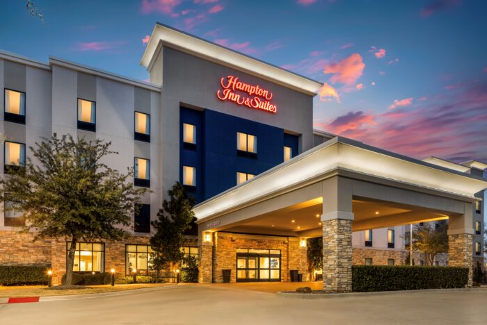A Hampton Inn & Suites hotel exterior at dusk, featuring illuminated Hampton Inn & Suites signage, a covered entrance, trees, and a vibrant sky with clouds in the background.