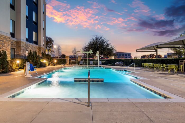 A lit outdoor swimming pool at dusk, surrounded by lounge chairs, tables, and umbrellas at Hampton Inn & Suites, next to a modern building with a colorful sunset sky in the background.