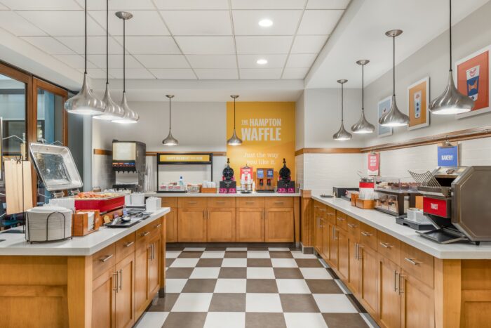 The Hampton Inn & Suites breakfast buffet area features a checkered floor, wooden cabinets, waffle makers, juice dispenser, baked goods, and cereal stations. A sign reads “The Hampton Waffle” beneath contemporary lighting overhead.