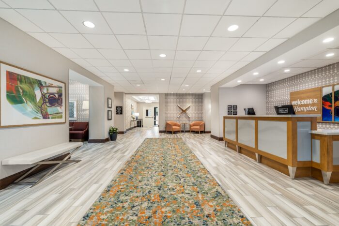 Hotel lobby at Hampton Inn & Suites with a patterned carpet runner, modern reception desk, two orange chairs, abstract artwork on the walls, and bright overhead lighting. A sign at the desk says Welcome to Hampton.