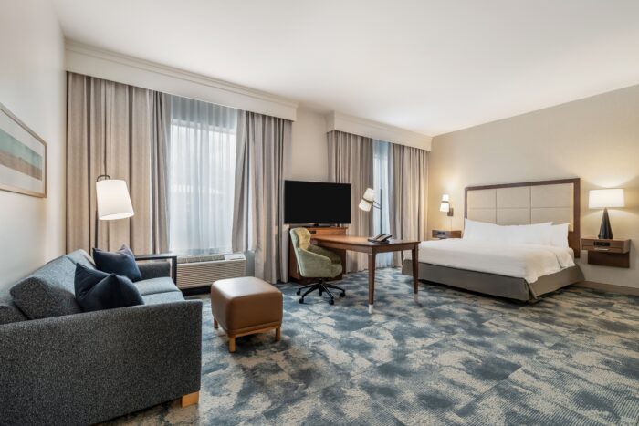 Modern Hampton Inn & Suites hotel room with a king bed, gray sofa, ottoman, desk with office chair, TV, lamps, and large windows with curtains letting in natural light. Light-colored walls and patterned carpeting add to the inviting feel.