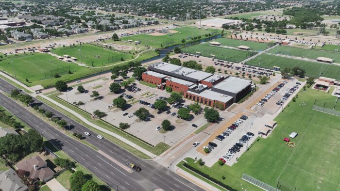 Aerial view of a large, red-brick building near Carpenter Park with a parking lot, green sports fields, roads, and surrounding neighborhoods on a sunny day. Many cars are parked around the building and fields.