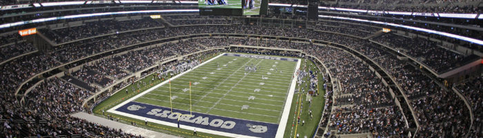 A wide-angle view of a packed football stadium with a game in progress on a field marked Cowboys, under a large video screen hanging from the domed roof. Spectators fill the stands on all sides.