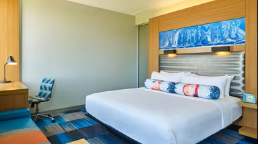 Modern Aloft Plano hotel room with a large bed, colorful pillows, artwork above the headboard, a desk with a lamp and chair, and patterned carpet. The decor is bright and contemporary.