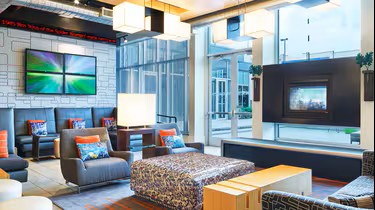 Modern hotel lobby at Aloft Plano with stylish seating, patterned ottoman, large windows, two TVs, and bright lighting. Blue and orange accents add color, while natural light fills the contemporary space.