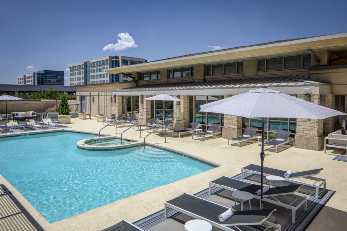 Outdoor pool area with lounge chairs, umbrellas, and a round hot tub. The modern Marriott Dallas/Plano building and other office buildings are visible in the background under a clear blue sky.