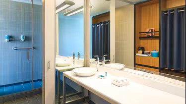 Modern bathroom at Aloft Plano with a large mirror, white countertop with sinks, towels, toiletries, and a glass-walled shower. Shelves with mugs and a blue ice bucket are visible in the background.