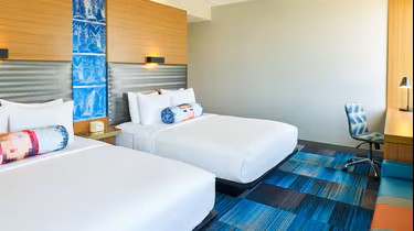 Experience comfort and style at Aloft Plano in this hotel room featuring two white double beds, colorful cylindrical pillows, a striped blue carpet, a modern desk chair, a desk with a lamp, and decorative wall art above the beds.