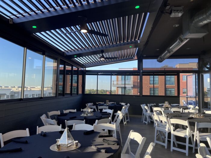 Rooftop Event Spot with white chairs and round tables draped in black cloths, set for a gathering. Large windows frame views of neighboring buildings, while a partly open slatted roof fills the space with natural light.