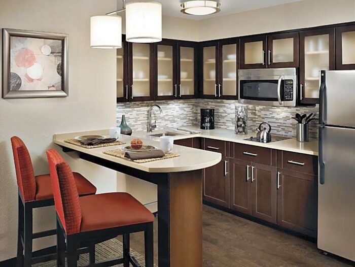 A modern kitchen at Staybridge Suites Plano featuring dark wood cabinets, a peninsula with two red bar stools, and stainless steel appliances.