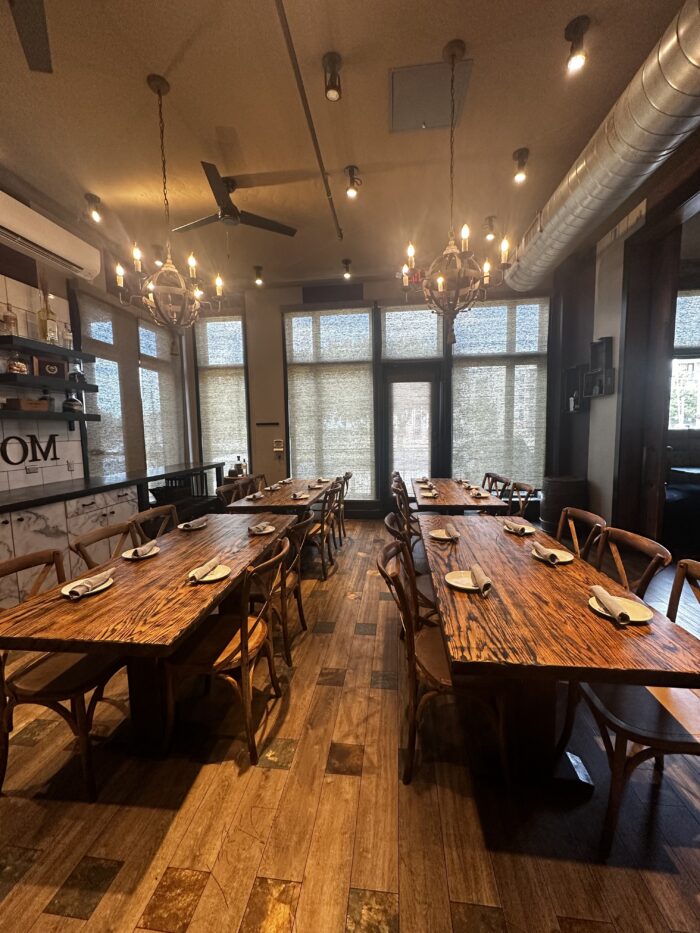 A warmly lit dining room at Urban Rio features two long wooden tables set with napkins, wooden chairs, rustic chandeliers overhead, wood floors, and large windows with blinds welcoming in natural light.