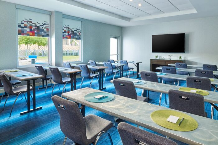 Modern classroom at Aloft Plano with rows of gray chairs and tables, notepads and pens on each desk, large TV on the front wall, windows with patterned shades, and blue striped carpet.