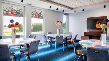 Modern meeting room at Aloft Plano featuring round tables, blue chairs, colorful flower centerpieces, a bright striped carpet, large windows with pixel-patterned blinds, and a wall-mounted TV.