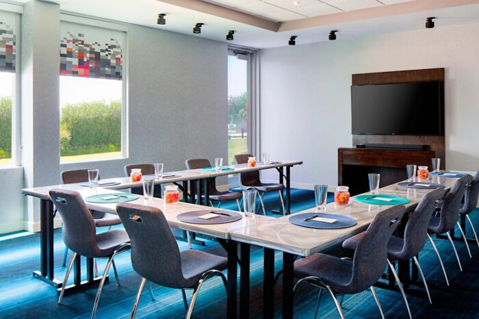 Modern conference room at Aloft Plano with two rectangular tables arranged in a U-shape, surrounded by gray chairs. Notepads, pens, glasses, and bowls of candy are thoughtfully placed on the tables. Large windows flood the space with natural light, while a TV screen provides added functionality.