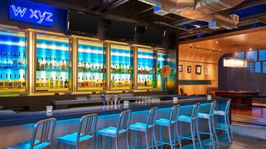 A modern bar at Aloft Plano features blue lighting, a neon w xyz sign, shelves of liquor bottles, metal barstools, and glasses neatly arranged on the countertop.
