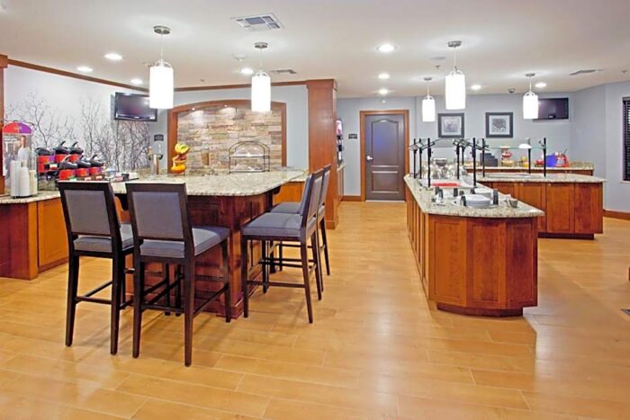 The breakfast area at Staybridge Suites Plano features a stone-accented counter, high-top bar stools, and buffet service stations with wood cabinetry.