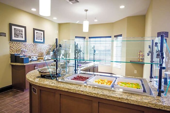 A hotel breakfast buffet station at Staybridge Suites Plano featuring three square bowls of raspberries, melon, and fruit salad behind a glass sneeze guard.