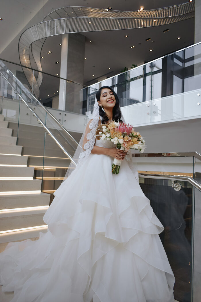 A bride in a flowing white gown and lace veil stands on a modern staircase at Renaissance Dallas, smiling and holding a colorful bouquet of flowers in a bright, elegant indoor setting.
