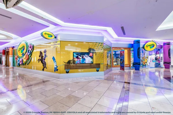 A bright, modern mall storefront for Crayola Experience Plano features colorful Crayola logos, playful decorations, and life-sized crayon characters on the windows. The shiny white tile floor completes the vibrant atmosphere.