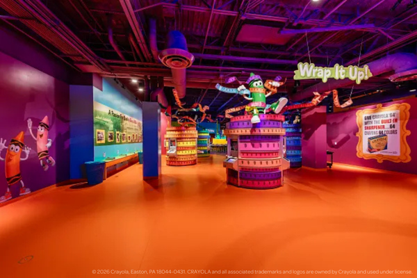 A colorful indoor play area at Crayola Experience Plano features bright orange floors, purple walls, cartoon crayon characters, and a “Wrap It Up!” sign above a central display. The vibrant space is inviting for children.
