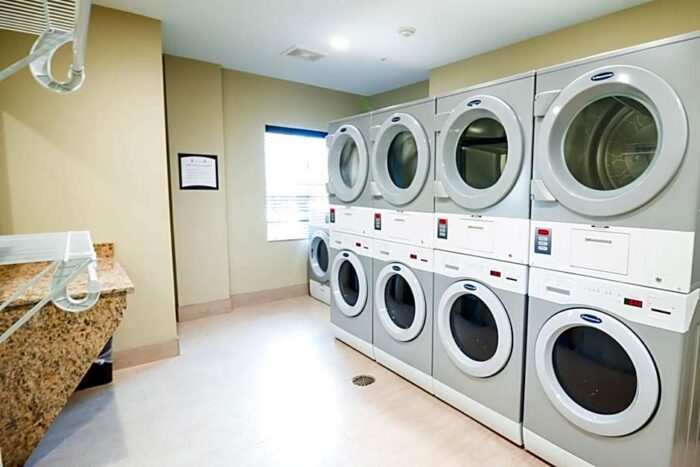 The laundry room at Staybridge Suites Plano features three vertical columns of stacked front-loading washers and dryers next to a granite-topped folding table.