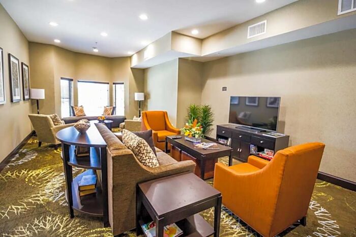 The Staybridge Suites Plano lobby features neutral-toned seating, two bright orange armchairs, a television on a media console, and a patterned carpet.