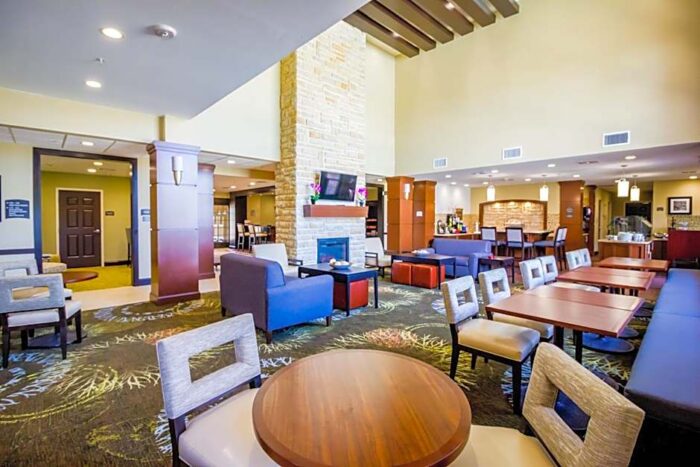A spacious Staybridge Suites Plano lobby featuring a central stone fireplace, lounge seating, and several dining tables and chairs.
