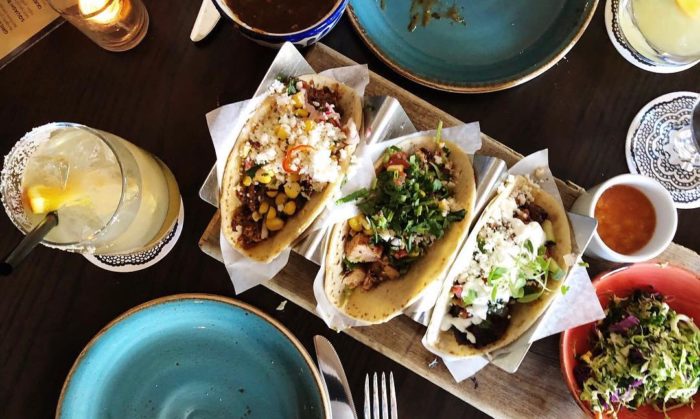 Three tacos filled with various toppings are served on a wooden board next to a green salad, a drink with a lemon wedge, sauce, and empty blue plates, all arranged on a dark table.