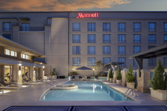 A hotel pool area at sunset with lounge chairs, umbrellas, and a hot tub, in front of the Marriott Dallas/Plano hotel building with lit windows and the red Marriott logo.
