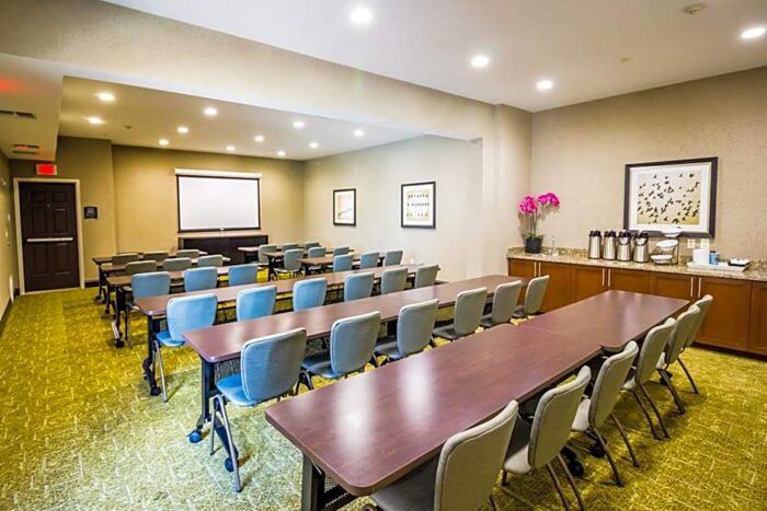 A Staybridge Suites Plano conference room arranged in a classroom setup with rows of tables and chairs facing a projection screen, and a beverage station along the back wall.