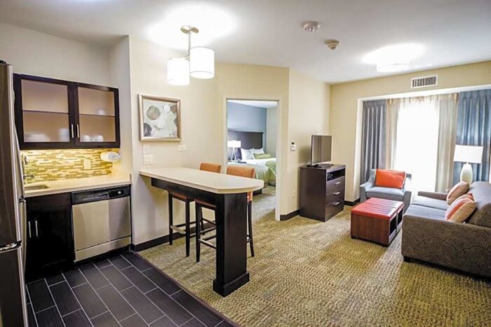 A Staybridge Suites Plano hotel suite featuring a kitchen with a bar counter, a living area with a sofa and television, and a partial view into a bedroom.
