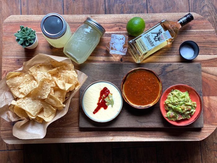 A platter with tortilla chips, queso dip, salsa, and guacamole sits on a wooden board. Nearby are a bottle of tequila, a lime, two jars with drinks, a small succulent, salt, and ice cubes on a wooden table.