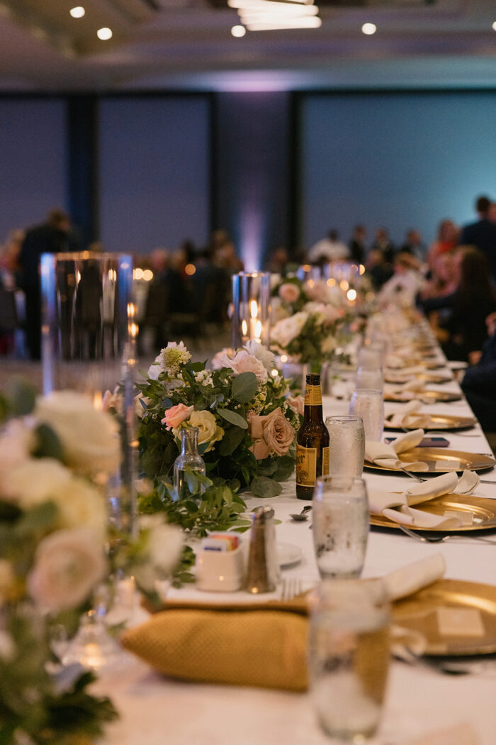 A long, elegantly set banquet table at Marriott at Legacy Town Center is decorated with white and pink flowers, candles, and gold plates; glasses, napkins, and drinks await as people are seated in the softly lit background.