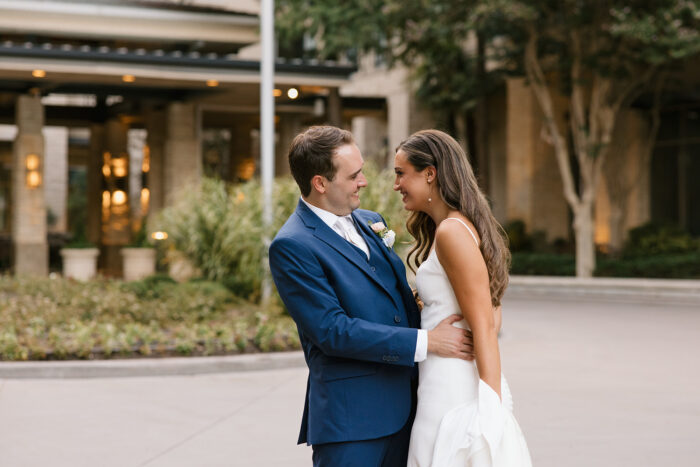 A bride and groom stand outside the Marriott at Legacy Town Center, smiling and embracing each other. The groom wears a blue suit and the bride holds her white dress in hand, framed by lush greenery and a building in the background.