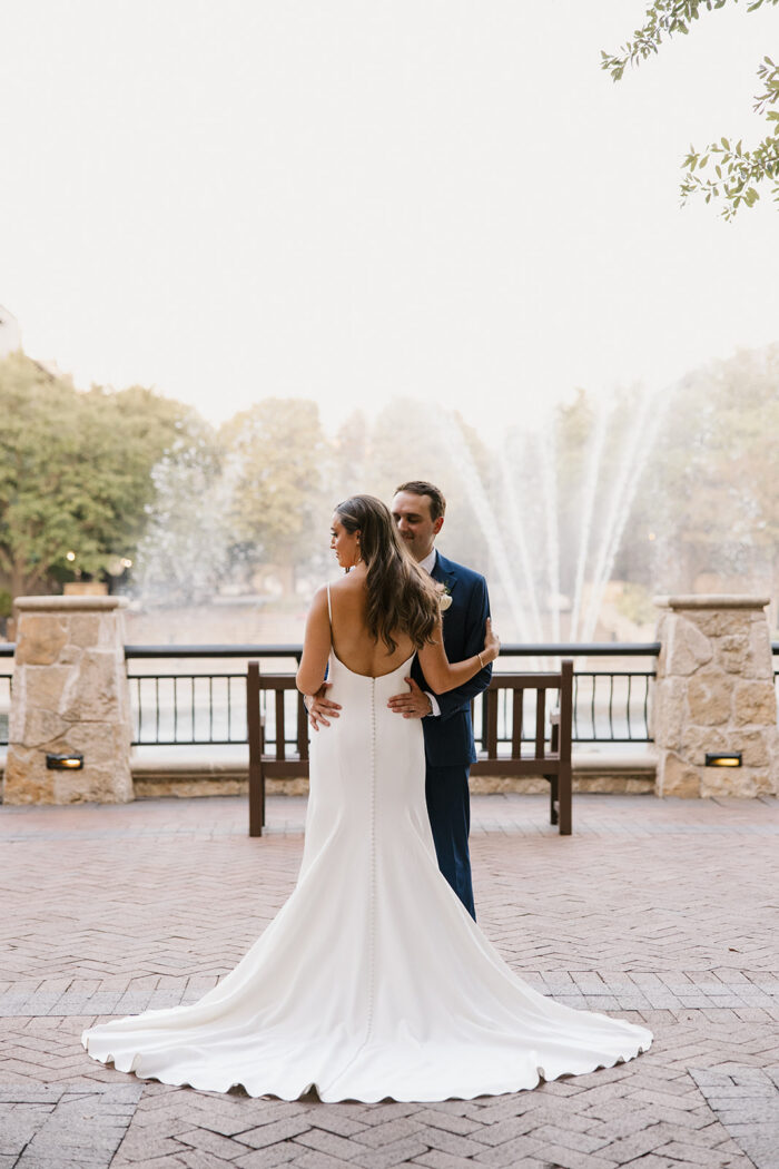 A bride in a white gown and a groom in a navy suit stand facing each other outdoors by a stone railing at the Marriott at Legacy Town Center, with a large fountain and trees in the background.