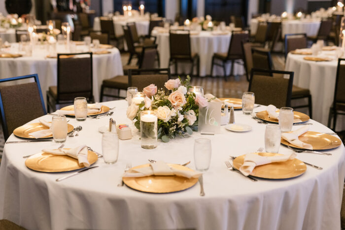 A round banquet table at Marriott at Legacy Town Center set for six with gold charger plates, white napkins, glassware, and a floral centerpiece of pale pink and white flowers. Other similarly set tables can be seen in the background.