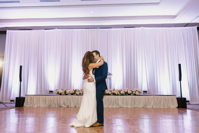 A bride and groom share their first dance in a spacious, elegant ballroom at the Marriott at Legacy Town Center, surrounded by white drapes, floral arrangements, and soft lighting as they embrace on the polished wooden dance floor.